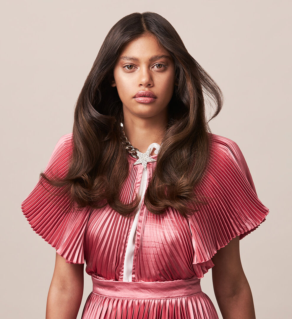 Portrait of a woman with long brown hair, wearing a pleated pink blouse with silver jewelry.